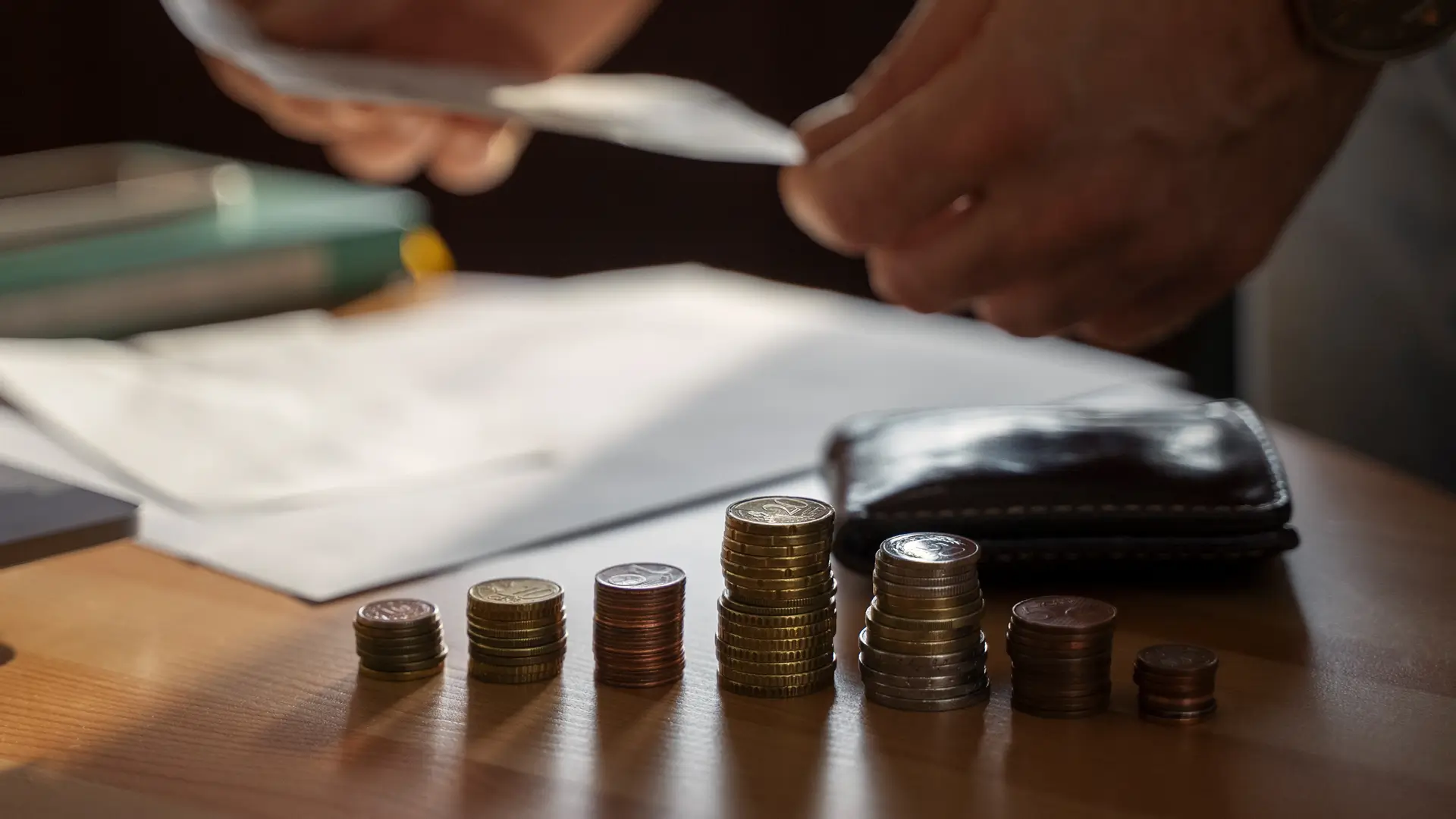 Pilha de moedas de diferentes tamanhos e valores colocadas em ordem crescente, com uma carteira e documentos ao fundo, representando economia e finanças.