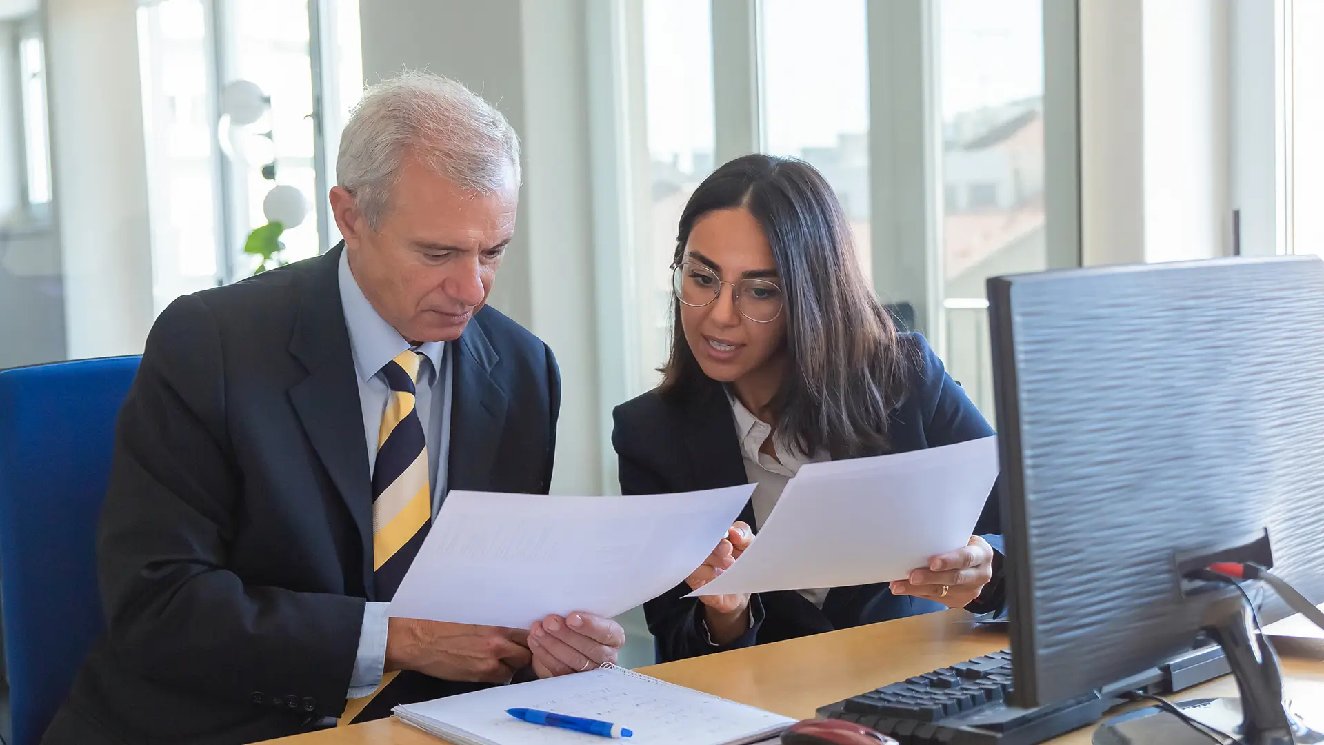 Homem de negócios e mulher analisando documentos tributários em ambiente de escritório, com computador na mesa, discutindo estratégias e planejamentos financeiros.