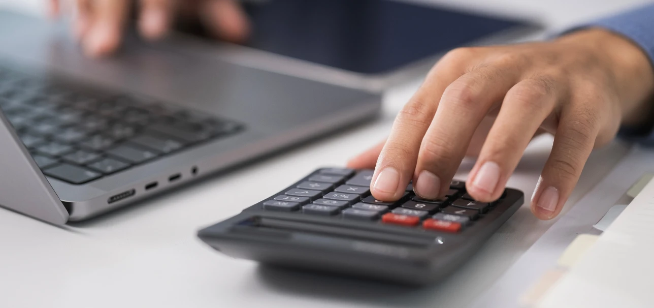 Pessoa trabalhando com calculadora e laptop na mesa, simbolizando análise financeira e cálculos precisos.